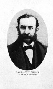 Daniel Coit Gilman is photographed facing the camera with his gaze of screen. The photo is balck and white. His dark hair contrasts sharply with his pale skin. He is wearing a dark suit, white shirt, and dark bow tie. He has a bushy beard and mustache with thinning hair on top. 