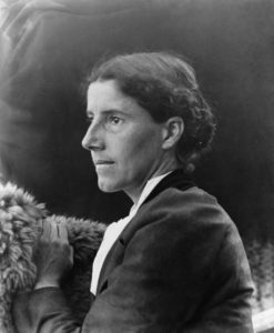 Charlotte Perkins Gilman faces off camera, her face in side profile. She wears a dark jacket with white shirt. Her hair is bound in a braid wrapped against her head. The photo is in black and white. 